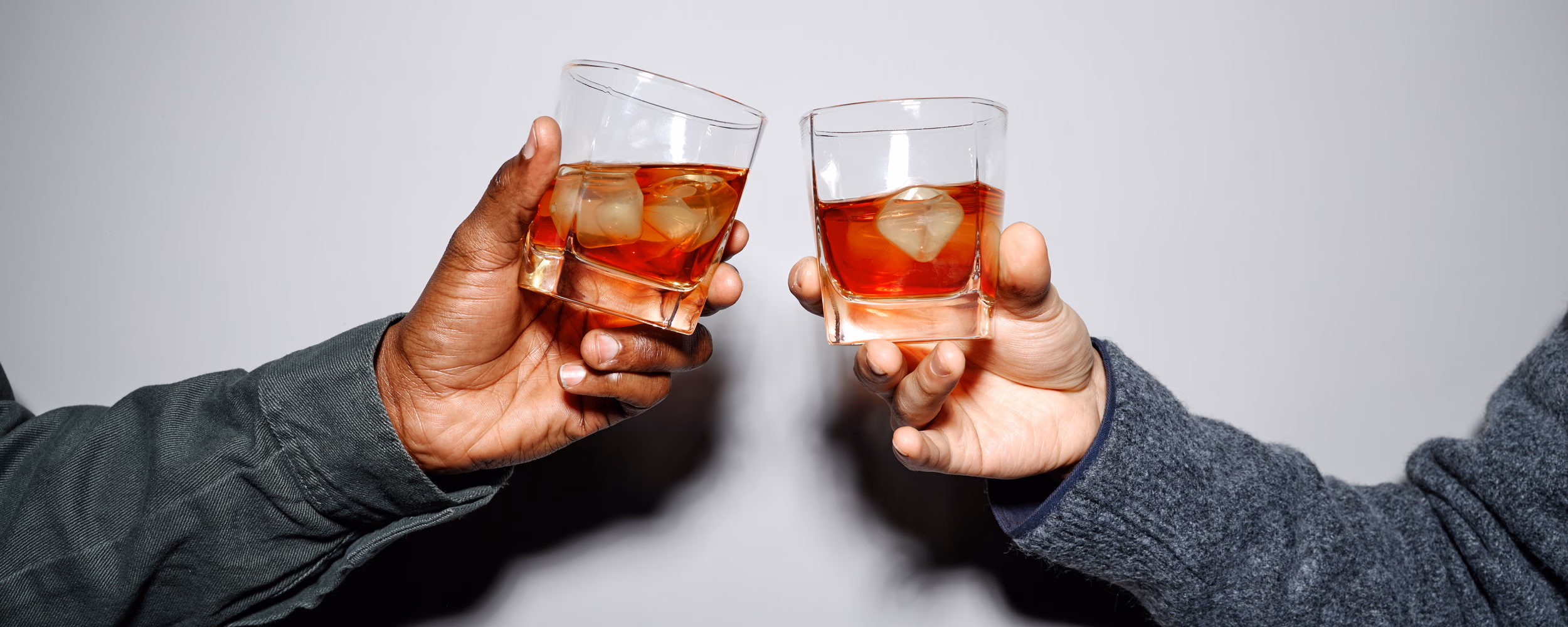 Close-up of two friends toasting with glasses of whiskey, highlighting camaraderie and celebration, with hands holding the glasses against a plain background